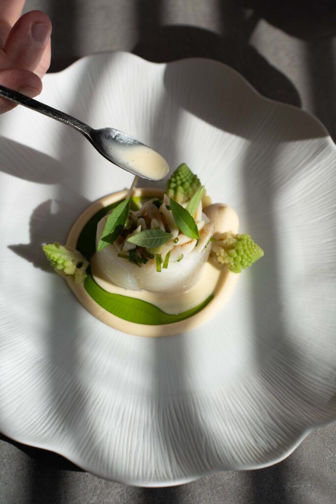 Chef schenkt saus over een visgerecht met romanesco en verbena op een golvend wit bord